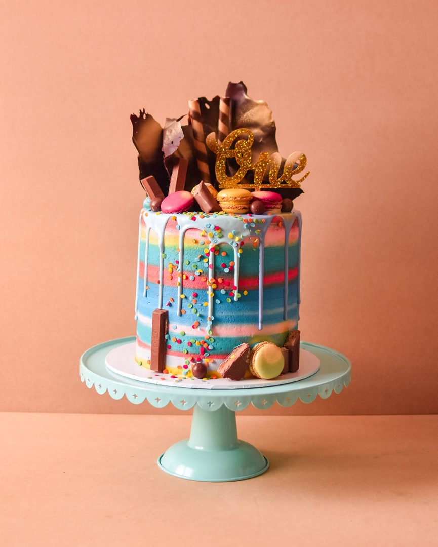 a rainbow cake with multiple colours sand chocolates and drip on top